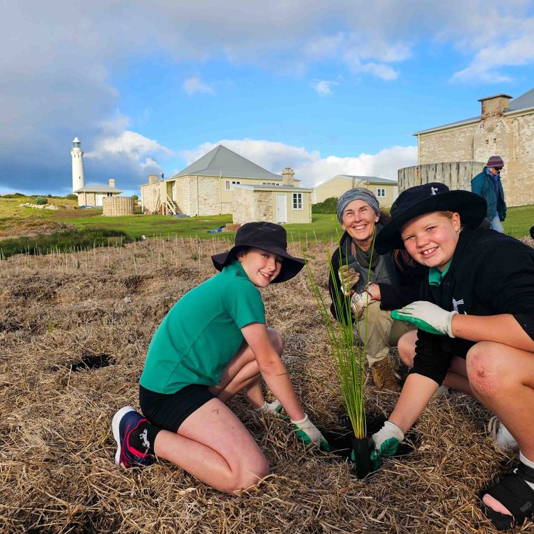 Group of people smiling while planting seedlings at Cape Leeuwin Lighthouse, with the lighthouse in the background.