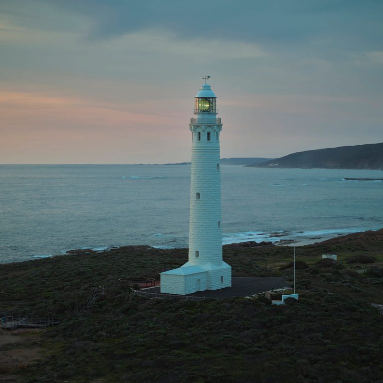 Cape Leeuwin Lighthouse glowing at sunset, casting its beam over the rugged coastline and sparkling ocean.