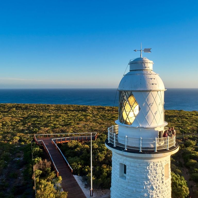 Three people on the Cape Naturaliste Lighthouse balcony, with sunlight streaming through the lighthouse lens and a view of the ocean beyond.