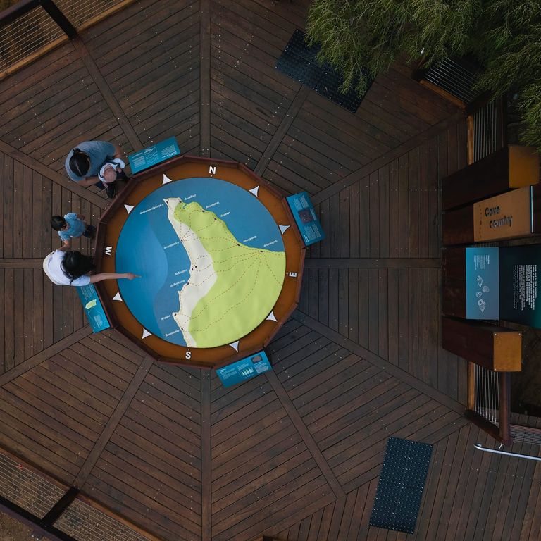 Aerial view of a family standing on a timber viewing deck at Ngilgi Cave’s Ancient Lands Experience, gathered around a large circular map of Cape Naturaliste with interpretive panels and bushland surrounding the platform.