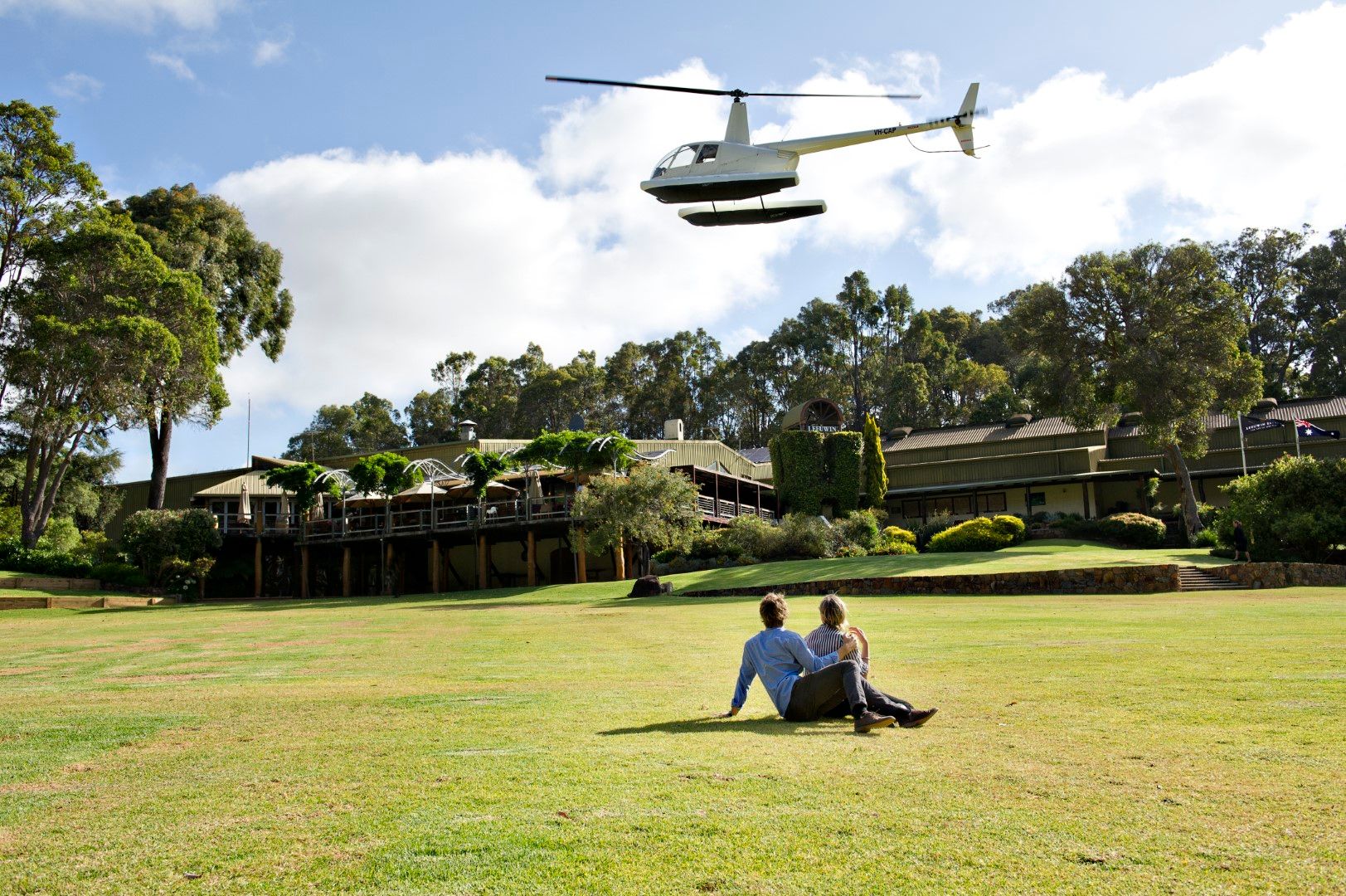 Couple sitting on the lawn at Leeuwin Estate while a helicopter flies overhead. Credit Scenic Helicopters.