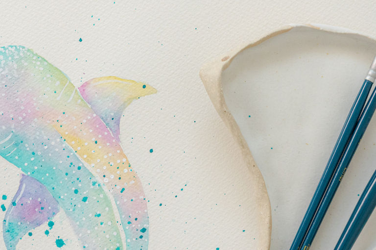 Aeriel view of three paintbrushes and a watercolour paint dish alongside a pastel coloured whale painting