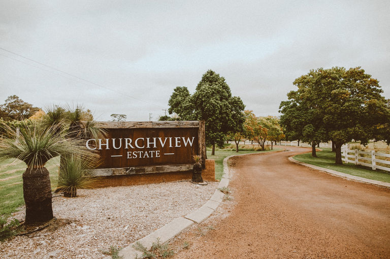 A dirt road leading into a winery with a brown 'Churchview Estate' sign on the left hand side