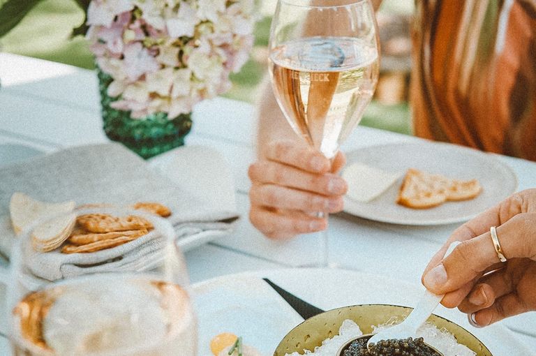 Close up image of a table with dips, crackers and cheese and wine, with two hands in the frame holding a wine glass and using a spoon