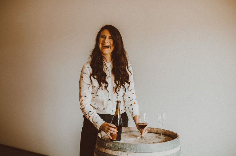 Woman with long brown hair in a white shirt looking to the left and laughing. She's holding a bottle of wine in her right hand and standing behind a wine barrel.