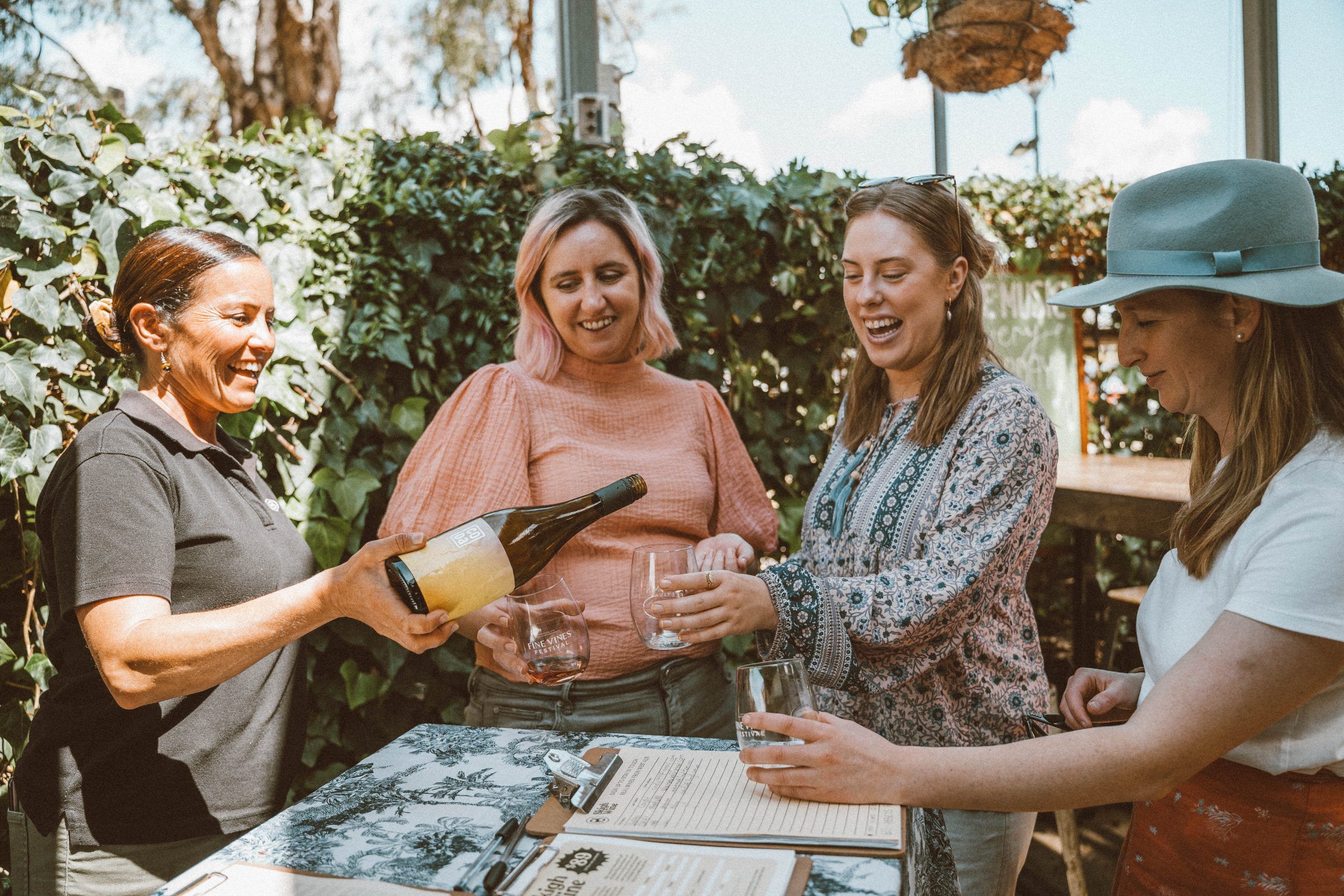 Four women standing around a table outside, the women to the left is pouring wine into their glasses