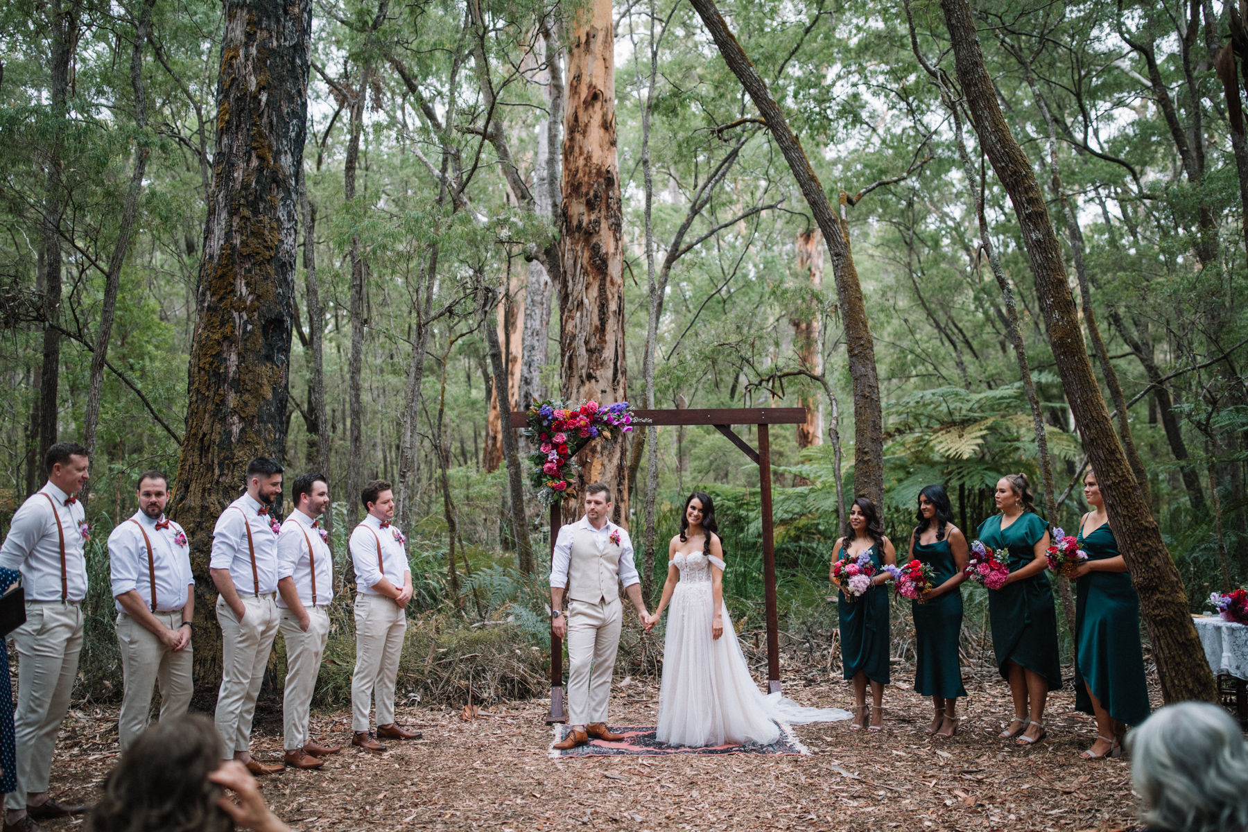 A wedding ceremony featuring bride and groom with 5 groomsmen in white shirts and grey pants and four bridesmaids in green dresses surrounded by tall trees in the forest