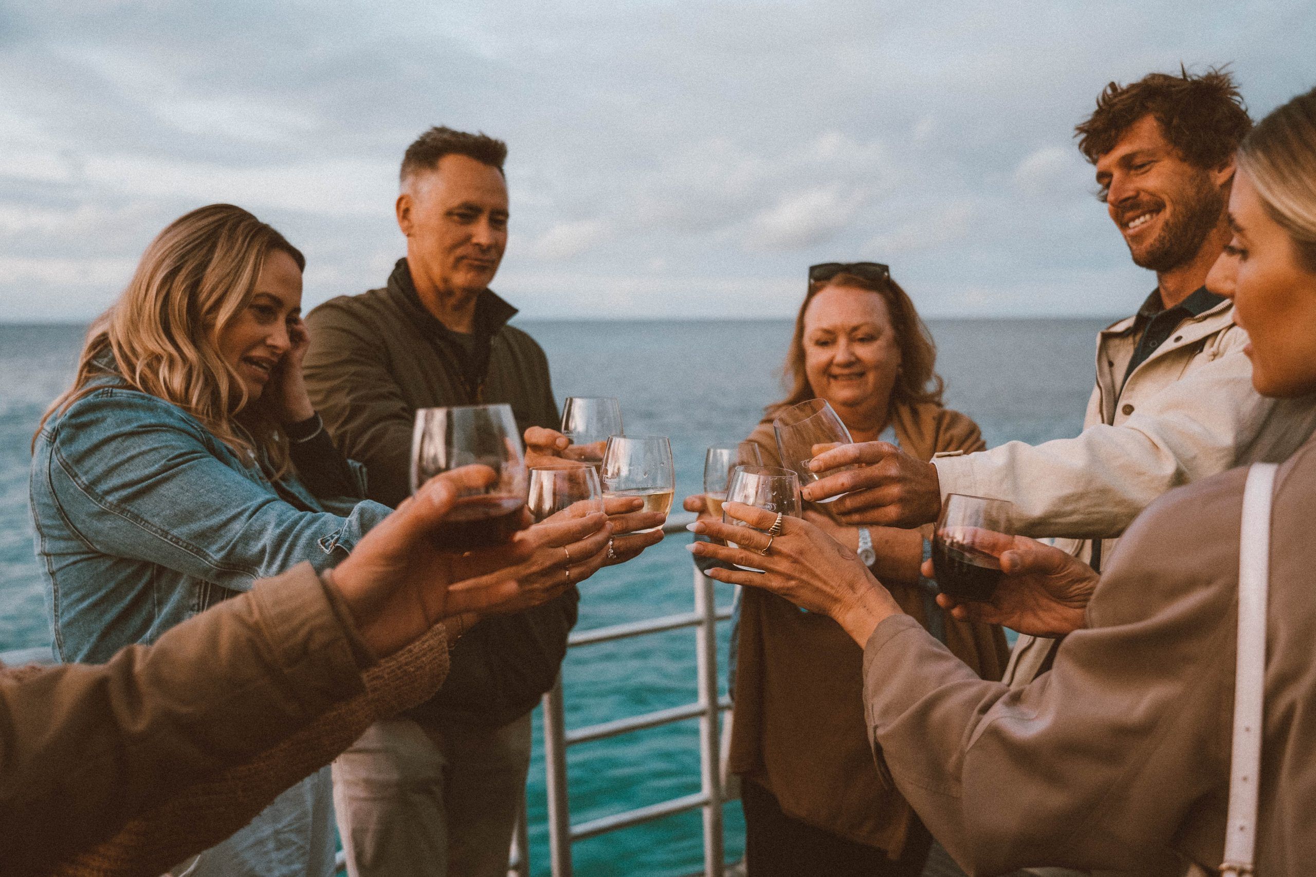 Image of 5 people, three women and two men, cheersing their glasses together on a boat with the ocean behind them