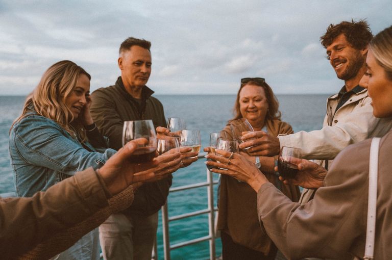 Image of 5 people, three women and two men, cheersing their glasses together on a boat with the ocean behind them