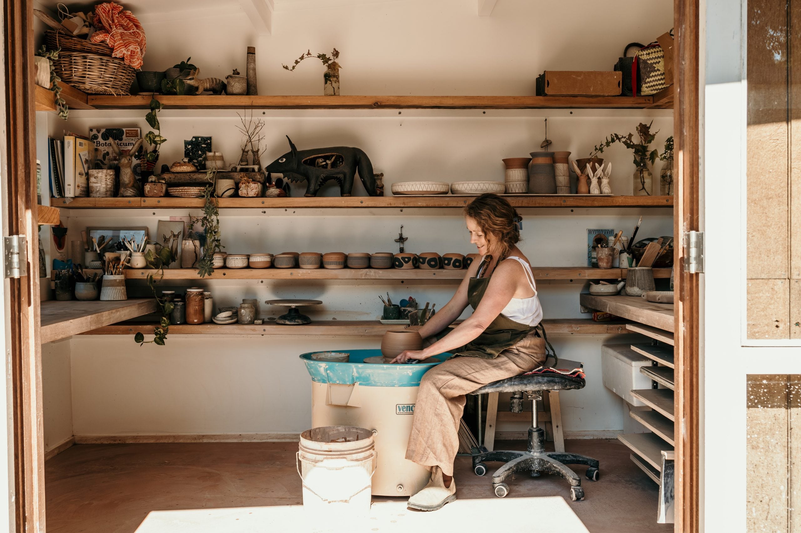 Jolene Hewison creating ceramic items inside their studio. There are several ceramic items, such as mugs, bowls and vases in the background.