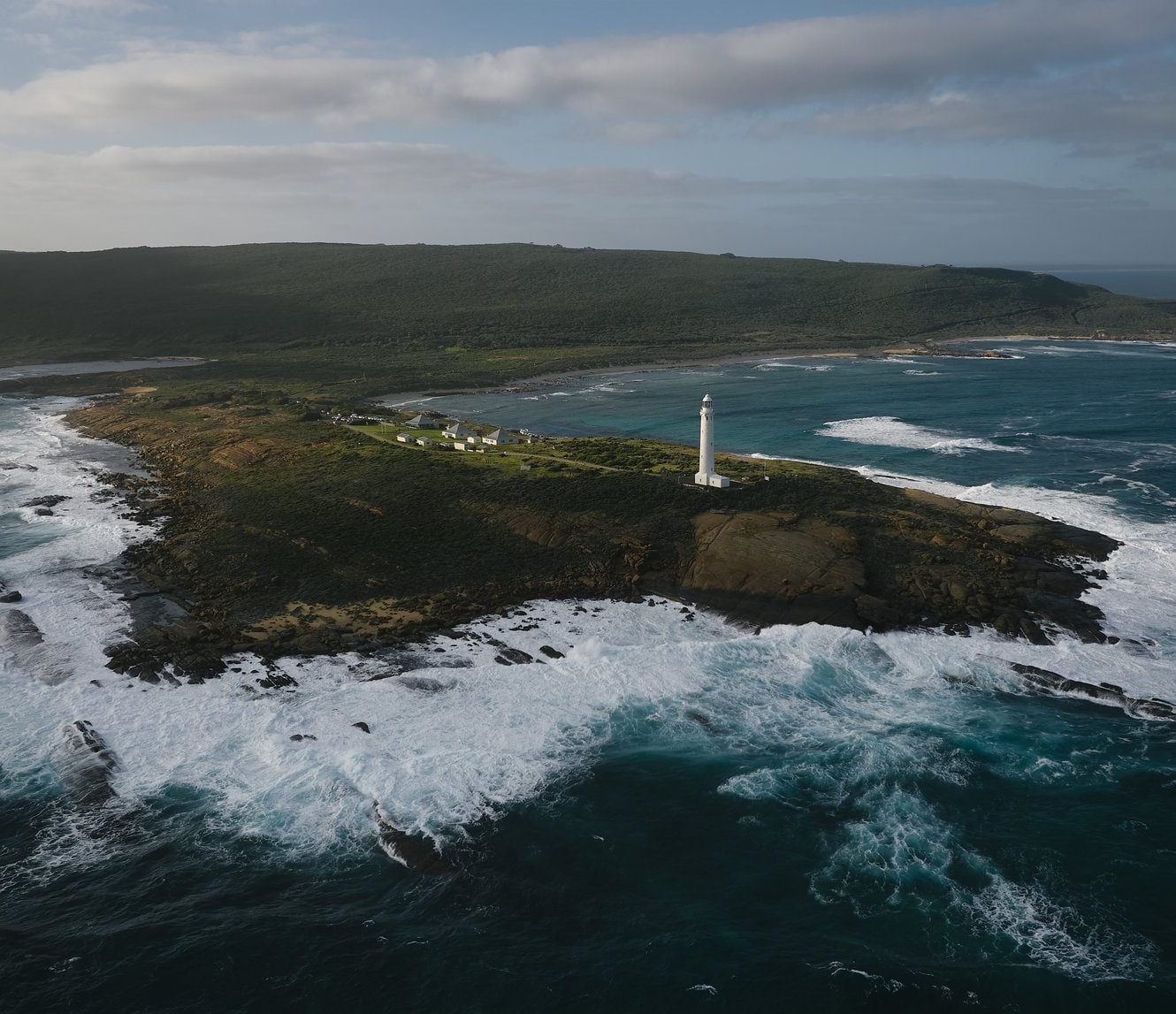 Aerial photo of Cape Leeuwin with the Cape Leeuwin Lighthouse in frame. Waves crashing on the rocks and deep blue ocean water