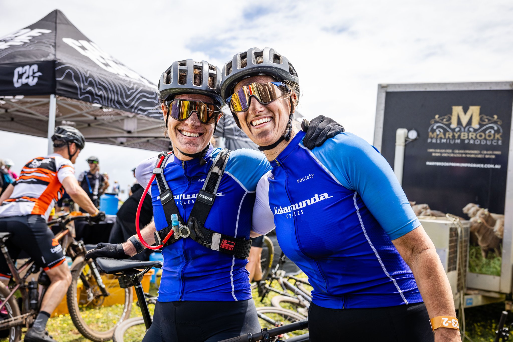 Two people standing next to each other smiling. Both people are wearing biking equipment, such as a helmet.