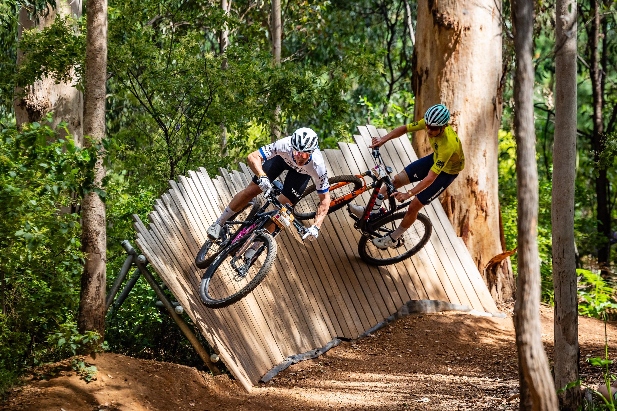 Two people riding mountain bikes over a wooden berm