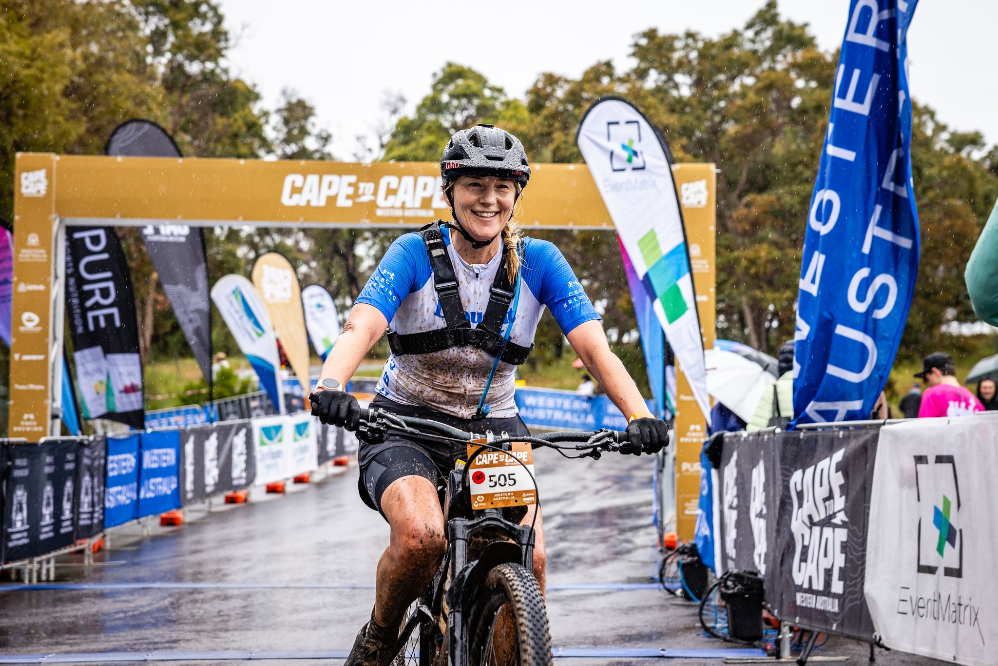 A person smiling while riding a mountain bike through a finish/starting line. There are signs and people in the background