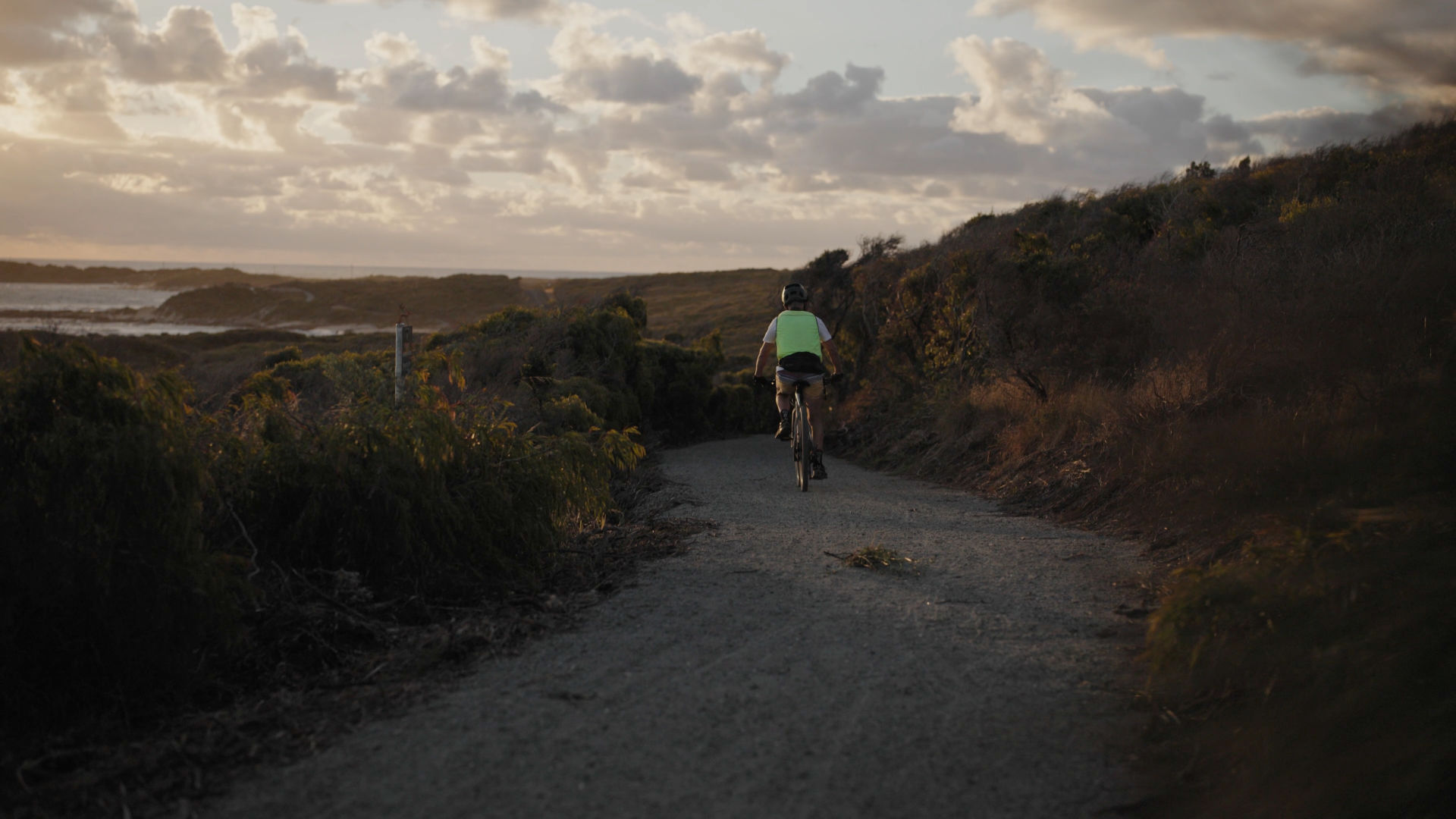 Photo of a man riding along a coastal gravel path at sunset.