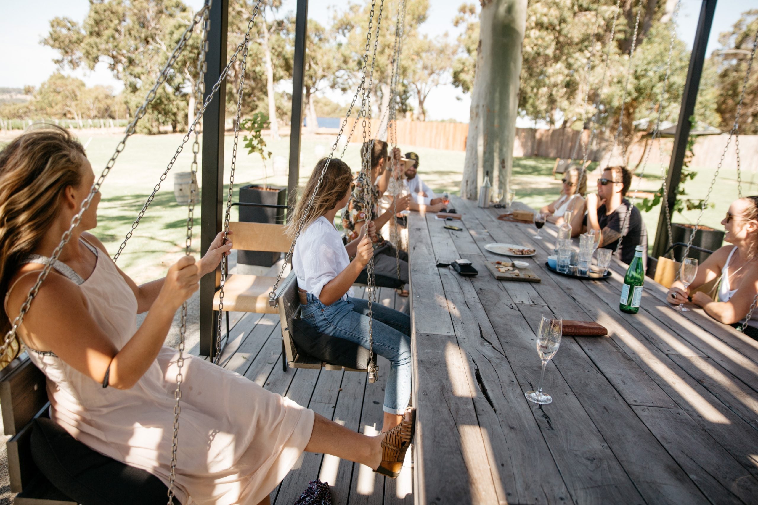 A group of friends relaxes around a long outdoor table at a Swings Yallingup, with several people seated on hanging swings and enjoying drinks in the sunshine.